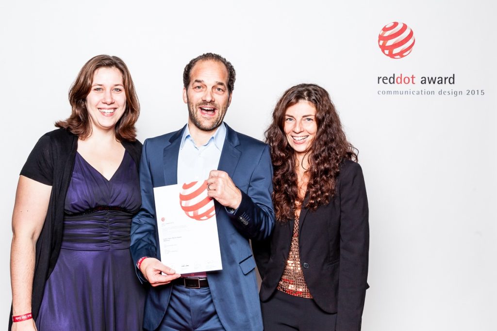 Drei Mitglieder des R211-Teams posieren mit einer Red Dot Award Urkunde für Communication Design 2015 vor einer weißen Fotowand