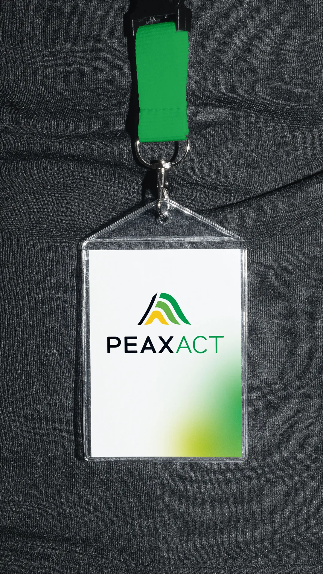 Name tag with green ribbon in front of a dark gray shirt, with the new PEAXACT logo with abstract wave motif in yellow, black and green to illustrate the Rebranding. Design and implementation by the R211 agency