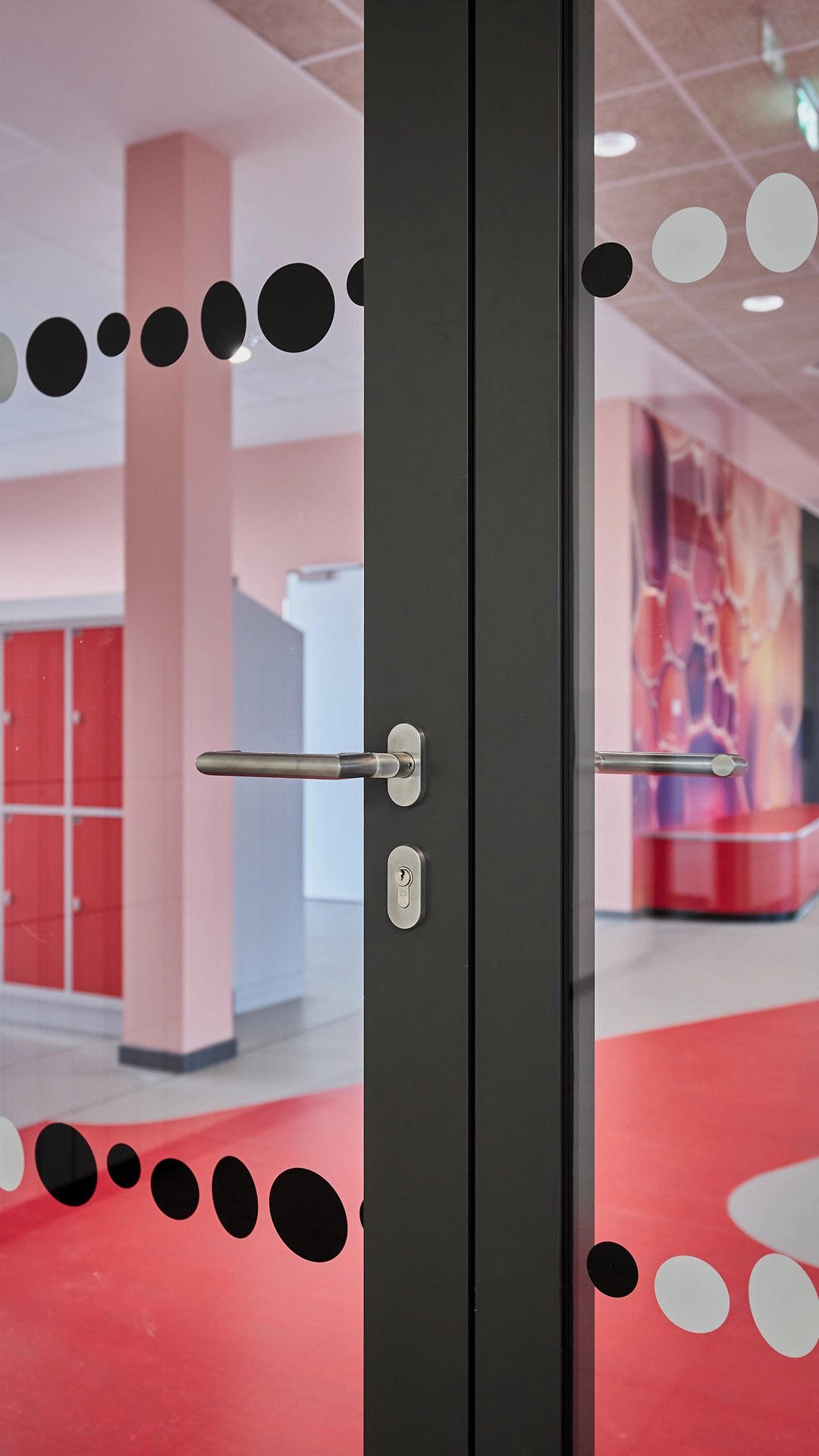 Schwarz-weißes Punktmuster auf Glastür als Bestandteil des Leitsystems von R211 in der Grundschule „Im Pfingsterfeld“, dahinter ein rot gestalteter Clusterbereich als Teil des farbcodierten Orientierungskonzepts
