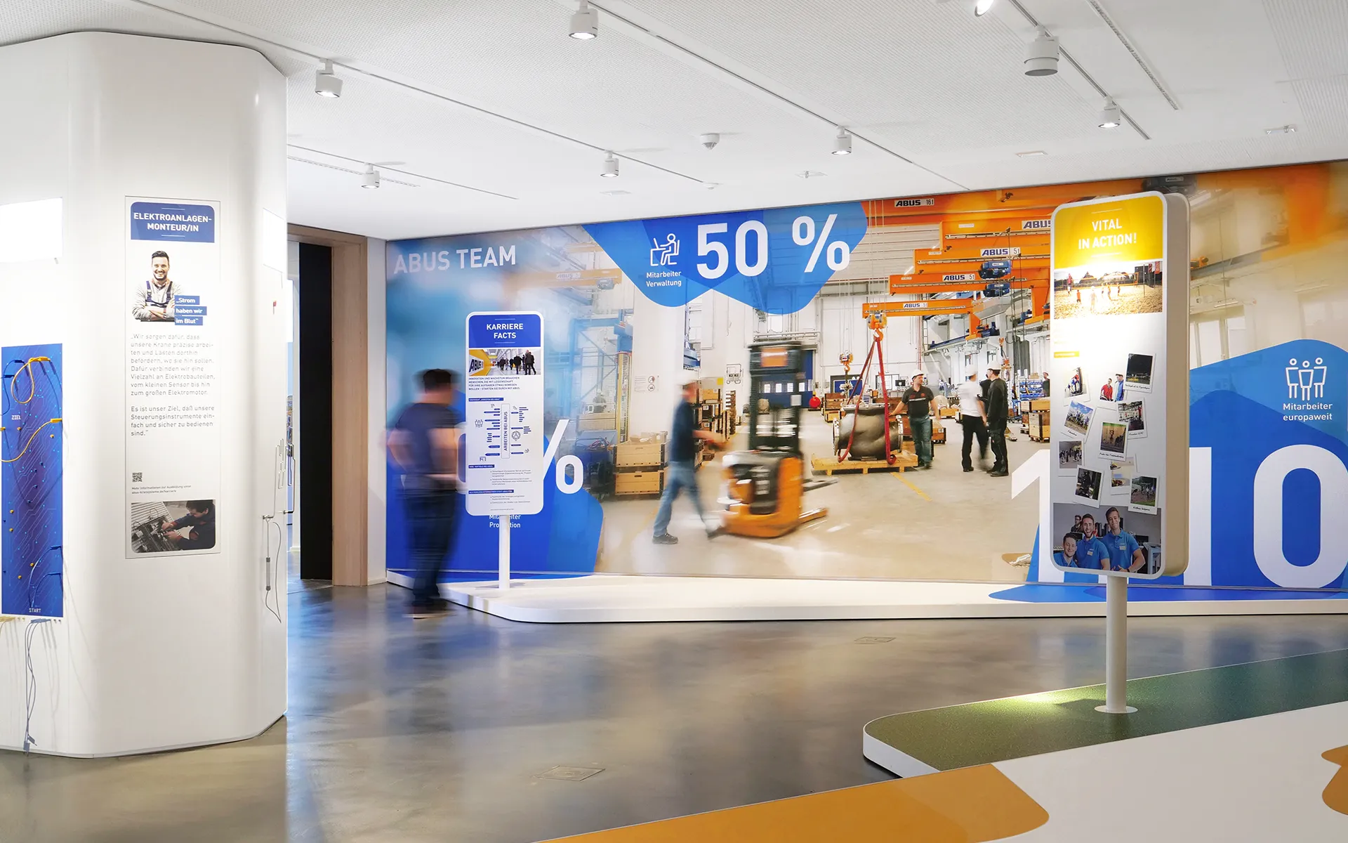Interactive exhibition and communication concept for a showroom for ABUS Crane Systems with large information walls, illuminated graphics, and visitors in motion.