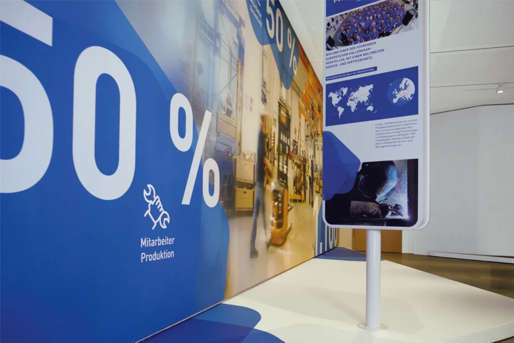Communication concept of a showroom for ABUS crane systems with an exhibition wall with blue design for production and info stele with world map and company data