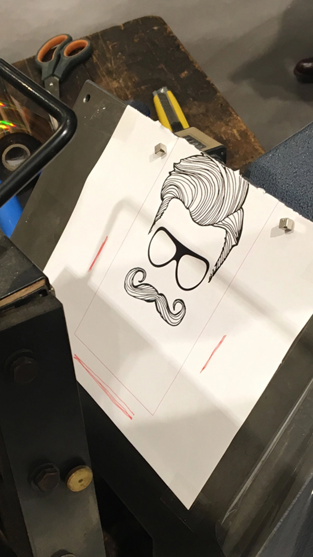 Papierbogen mit schwarz-weißer Illustration eines Mannes mit Haartolle, Brille und Schnurrbart, befestigt an einer Druckmaschine im Arbeitsprozess