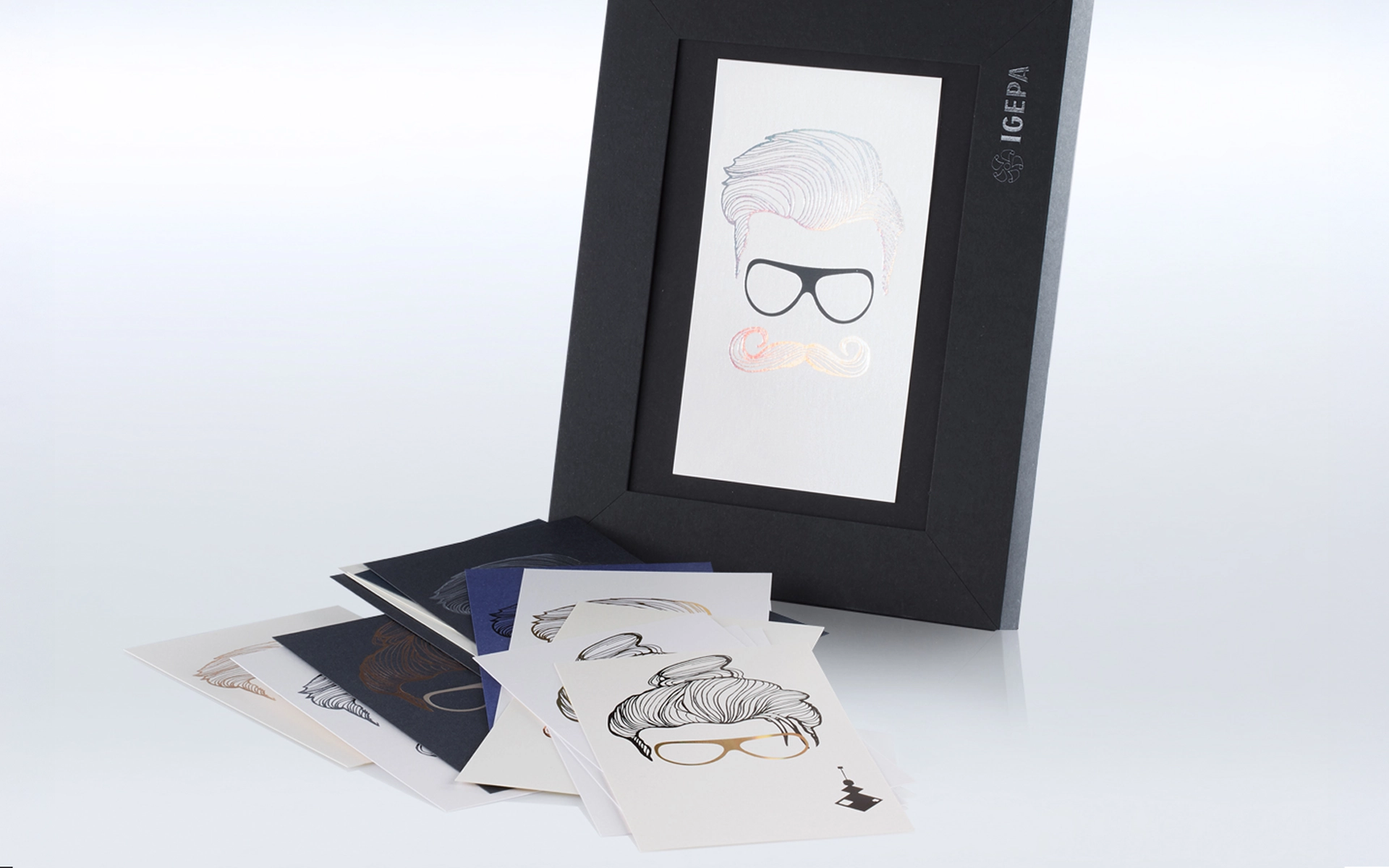 Mehrere Designkarten mit geprägten Illustrationen von Haaren, Bärten und Brillen in verschiedenen Metallicfarben, daneben schwarze Mappe mit IGEPA-Logo und eingelegter Karte mit stilisiertem Männerkopf. Aufnahme vom Paper Playground Event von Freytag & Petersen, visuell gestaltet von der R211-Agentur.