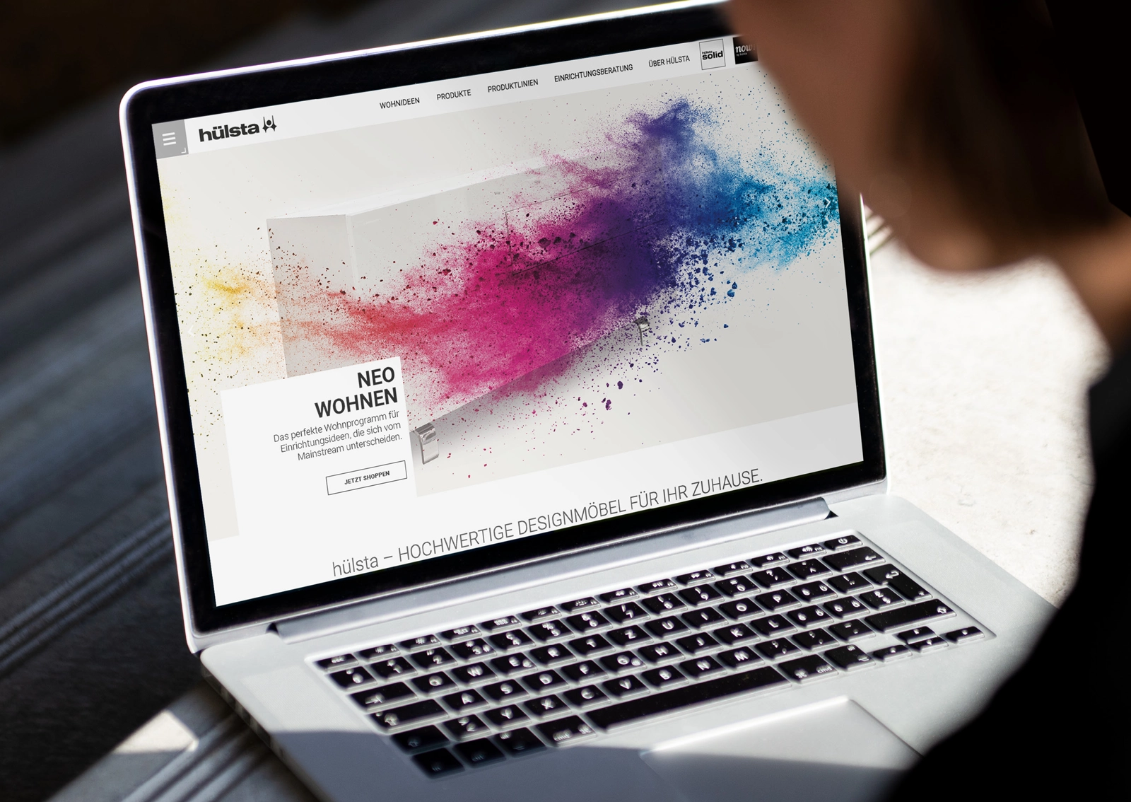 Laptopansicht der hülsta Website – Ankündigung eines Showroom-Events in Stadtlohn, visuell inszeniert mit weißem Möbelkubus und Farbexplosion in Magenta und Orange